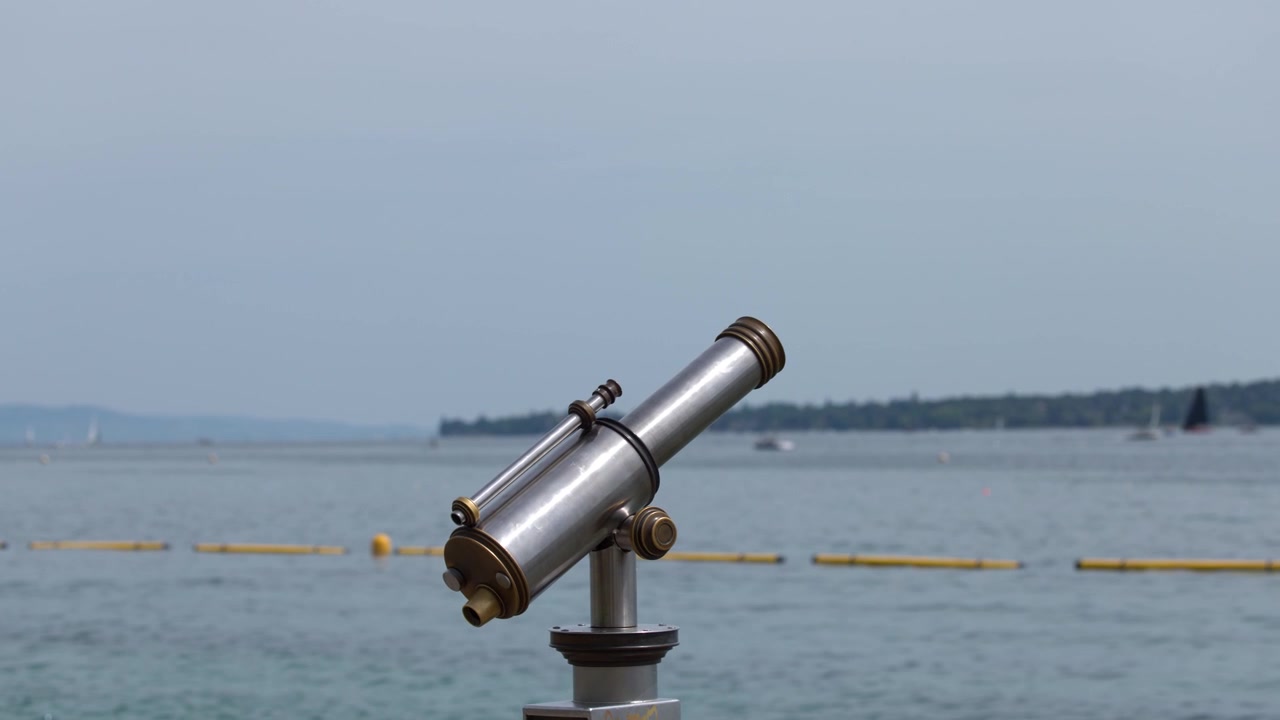 Static medium shot of a silver coin-operated telescope facing a calm lake under overcast skies.
