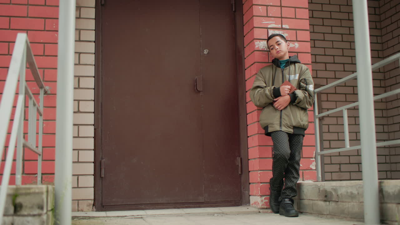 Kid in hooded jacket leans against red brick wall near building entrance, holding brown book while gazing thoughtfully into distance with calm and reflective expression