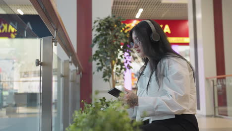 side view of lady in white shirt holding phone in right hand while operating it and squeezing stress ball in left hand, wearing headphones, standing indoors with plants and mall store in background
