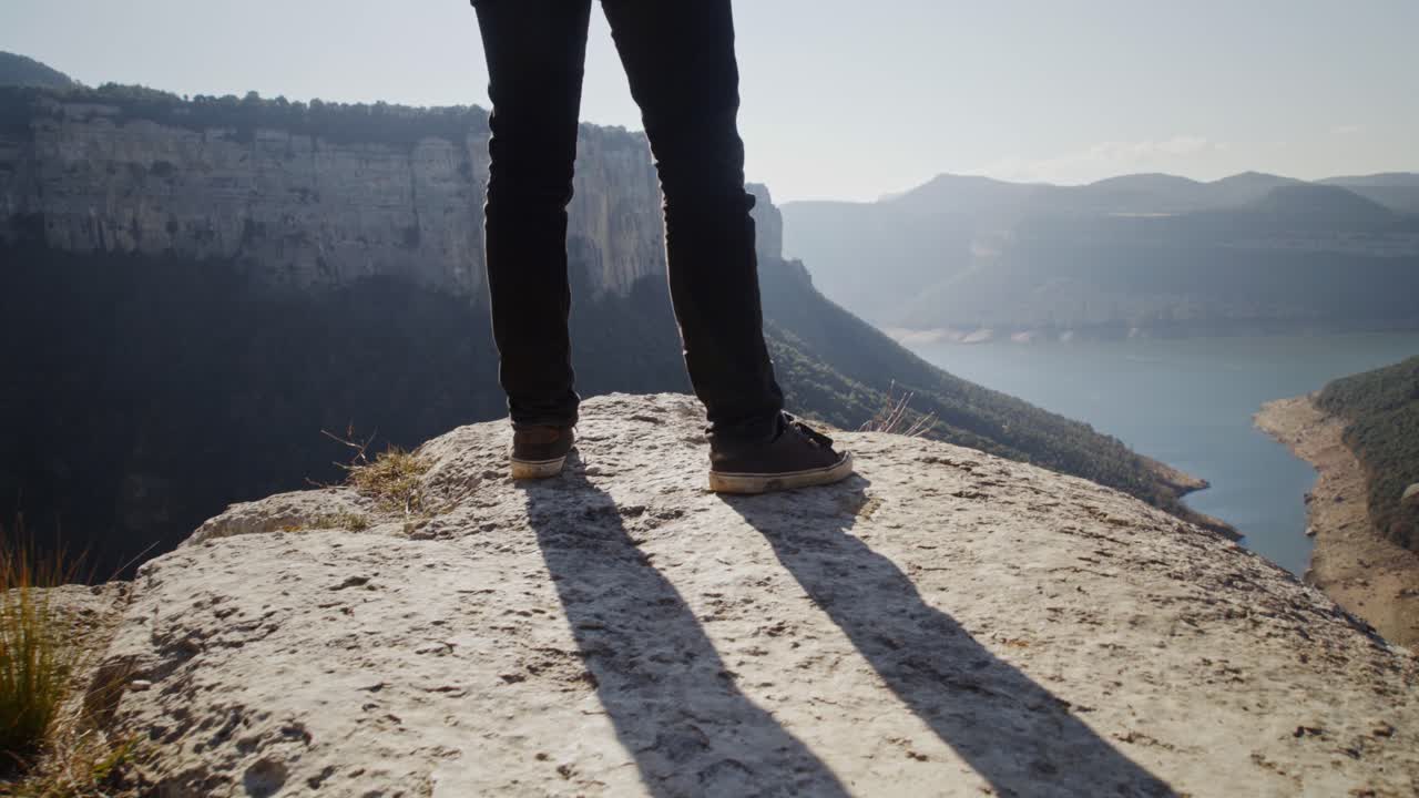 vista escénica de las cadenas montañosas y el lago en el fondo con una vista de ángulo bajo de las piernas del hombre de pie peligrosamente al borde del acantilado observando el paisaje, primer plano de mano