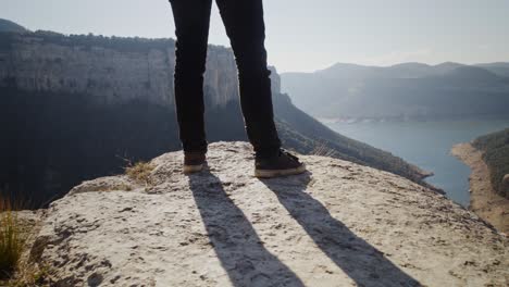 scenic view of mountain ranges and lake in background with close up low angle view of man's legs standing dangerously at edge of cliff observing landscape, handheld closeup