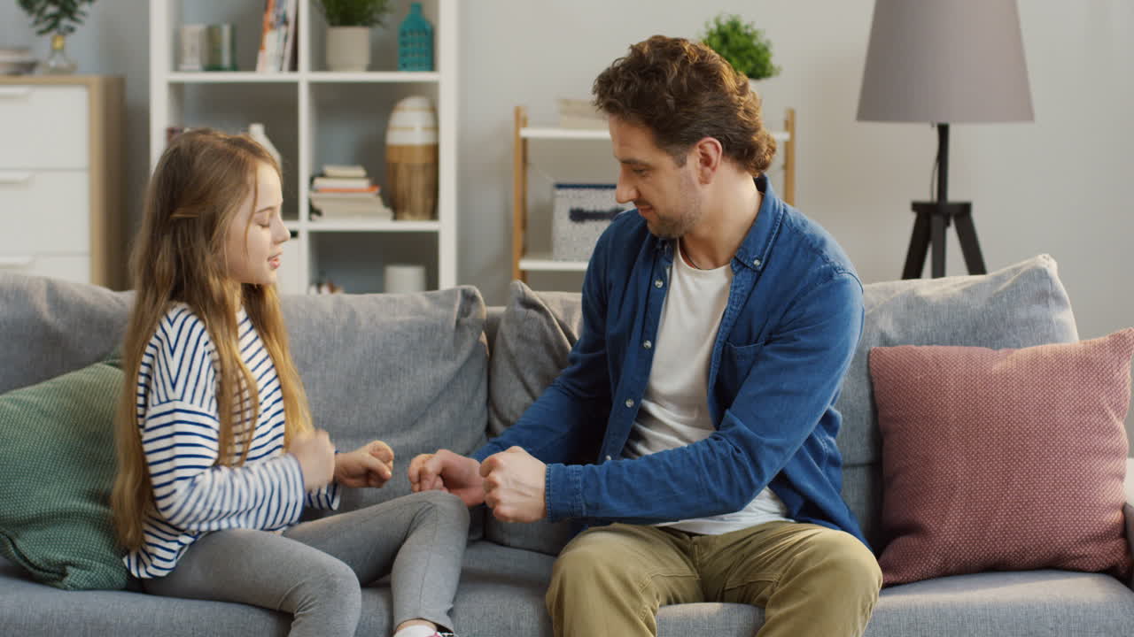Little Blonde Pretty Girl Sitting On The Couch With Her Father And They Playing A Hands Game