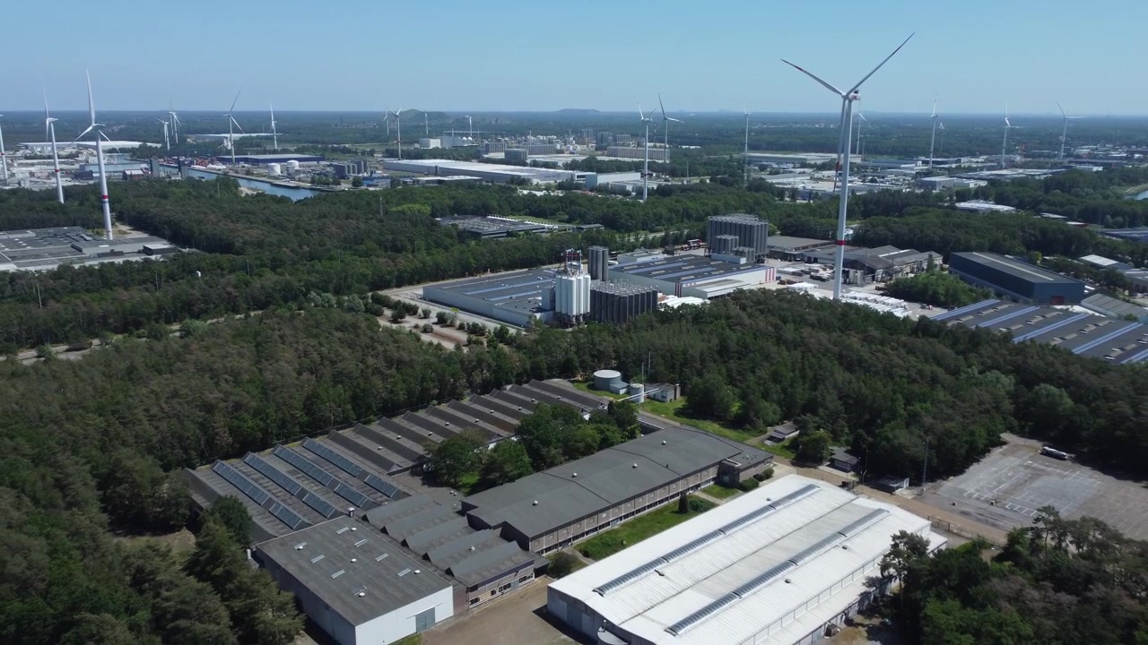 Drone footage flying out from an industrial estate, revealing storage silos, warehouses, scattered trees between the buildings, and wind turbines under a clear sunny sky.