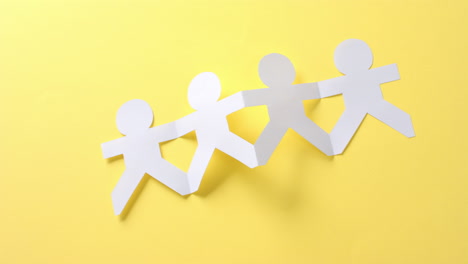 Close-up-of-people-holding-hands-made-of-white-paper-on-yellow-background-with-copy-space