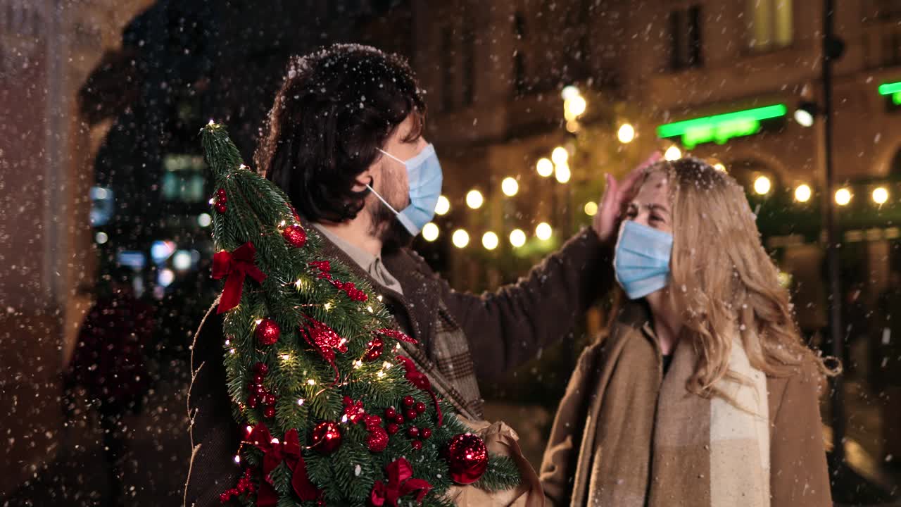크리스마스 트리를 들고 크리스마스 거리에서 눈이 내리는 동안 웃는 얼굴 마스크를 쓴 백인 행복한 커플