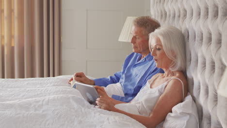 Pareja-De-Ancianos-En-Casa-En-La-Cama-Aislándose-Con-Tableta-Digital-Y-Leyendo-Durante-El-Cierre-De-Covid-19