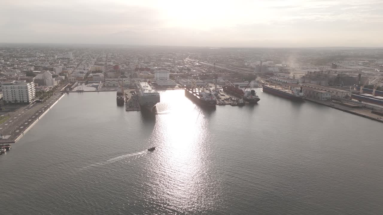 Aerial view of a busy seaport at sunrise, showcasing large cargo vessels, container terminals, industrial infrastructure, and an organized city layout in the background.