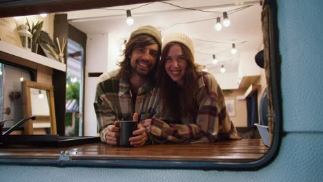 Portrait-of-a-happy-brunette-girl-in-a-white-hat-in-a-plaid-shirt-together-with-her-brunette-boyfriend-in-a-plaid-shirt-drinking-tea-in-a-trailer-and-relaxing-during-their-picnic-in-a-camp-outside-the-city-in-the-summer