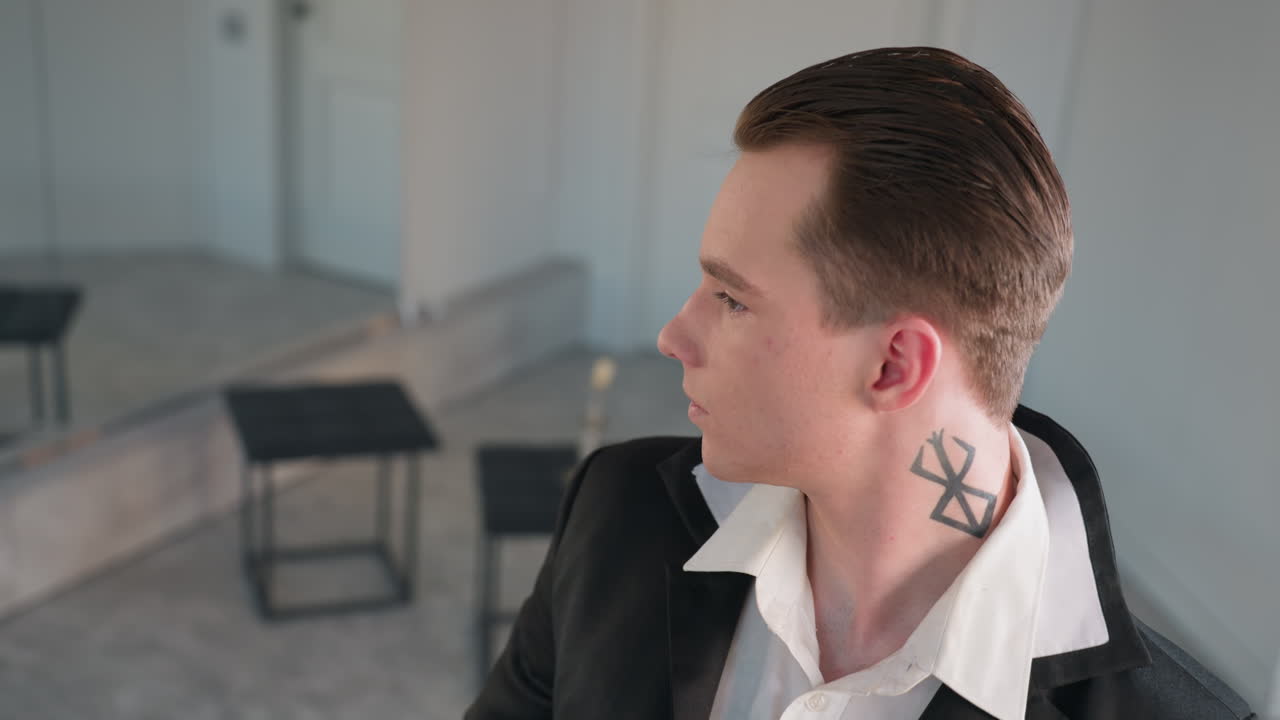man with neck tattoo and slicked hair looking elegant in black coat white shirt, three stools blurred behind, candle stand and mirror reflection, soft natural light on neutral studio walls