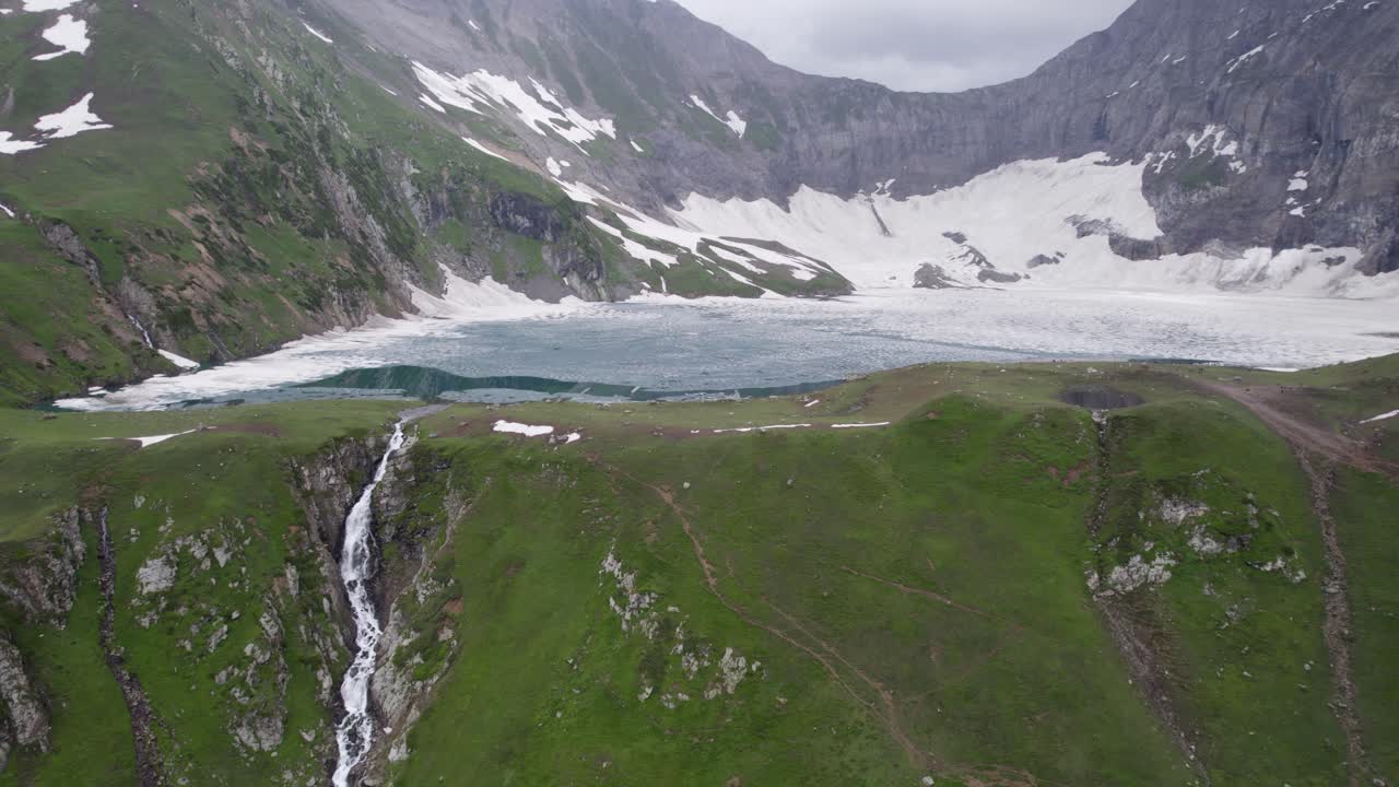 una cascada que desemboca en un lago alimentado por glaciares, rodeado de verdes prados y montañas cubiertas de nieve