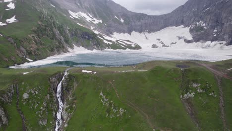 waterfall flowing into a glacier-fed lake, surrounded by green meadows and snow-covered mountains