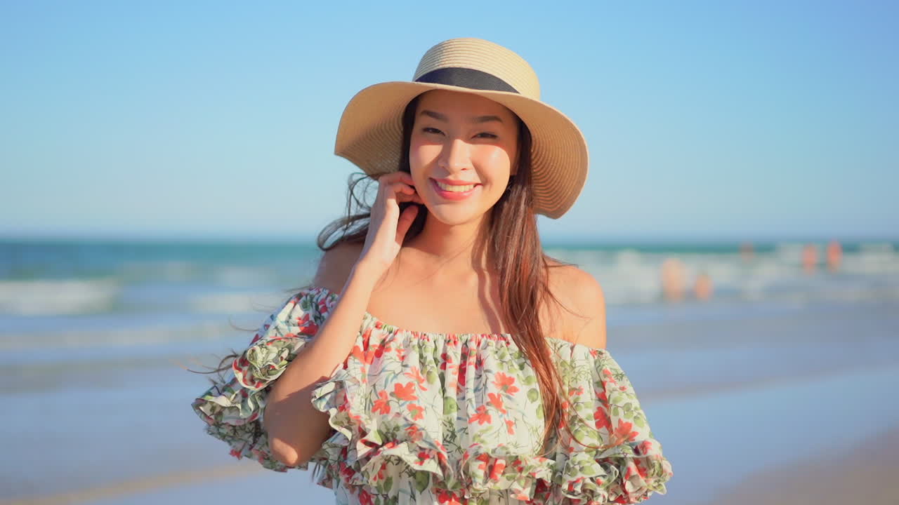 Beautiful Asian woman in frilly dress and straw hat standing beside the ocean. Breeze blows hair and dress of woman at the beach. Lady smiles and brushes hair from face at coast