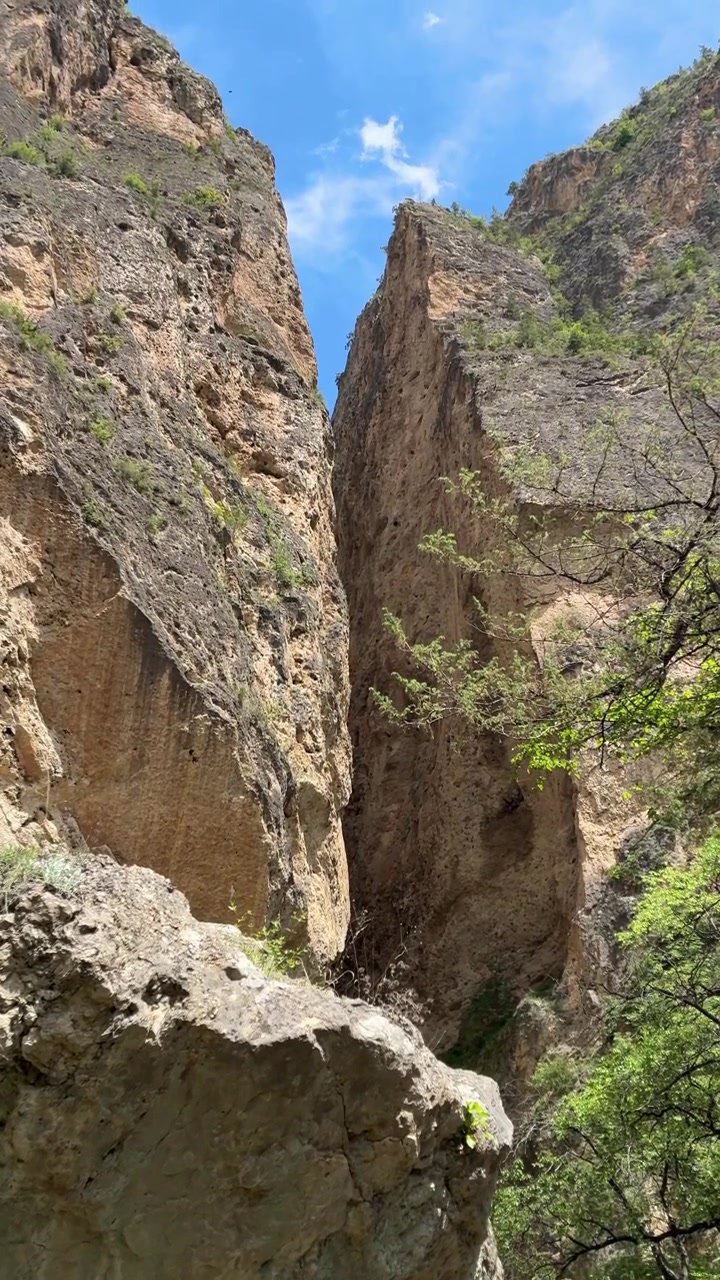 Una toma vertical en ángulo bajo captura un estrecho desfiladero de montaña con imponentes acantilados rocosos y árboles verdes bajo un cielo azul brillante
