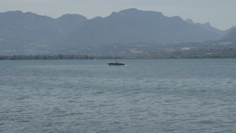 In-Der-Ferne-Segelt-Ein-Boot-Durch-Blaues-Wasser-Mit-Bergen-Und-Einer-Bewölkten-Stadt-Im-Hintergrund