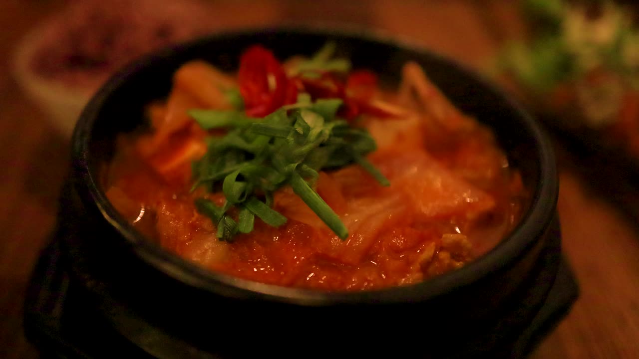 primer plano de sopa de kimchi burbujeante en una olla negra, adornada con verduras y chile rojo, bajo una iluminación cálida