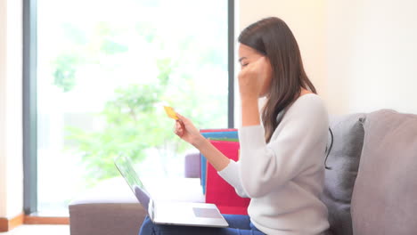 Mujer-Asiática-Sentada-En-El-Sofá-Con-Su-Laptop-En-El-Regazo-Y-Escribiendo-Tarjeta-De-Crédito-O-Número-De-Tarjeta-De-Descuento-Y-De-Repente-Mostrando-Una-Expresión-Ganadora-Y-Un-Gesto-De-Sí