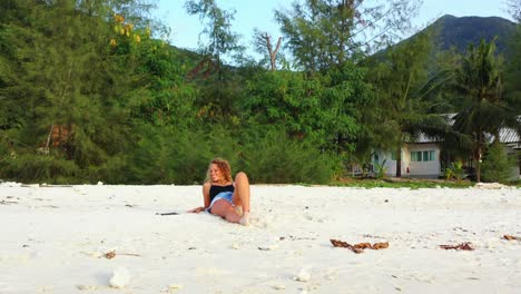 beautiful blonde girl lying on white sand of exotic beach relaxing under beautiful sunlight on tropical island with lush vegetation