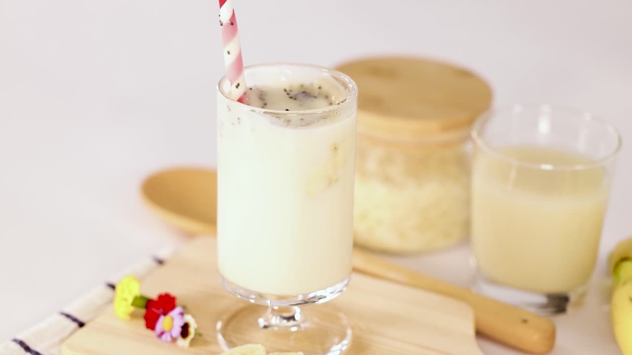 Hand stirs banana milkshake in glass, bright kitchen lighting, static camera, inviting healthy refreshment