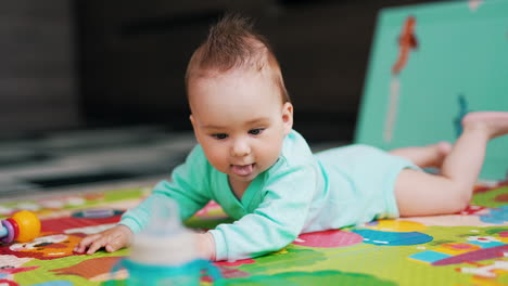 nice little caucasian toddler lies on the mat. baby pays attention to a bottle standing beside. blurred backdrop.
