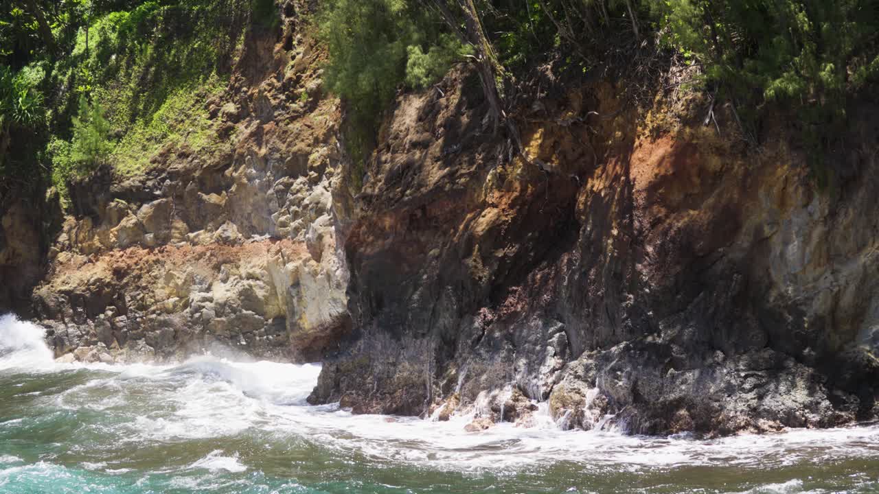 Powerful surf rolls in and detonates against a rough cliff face, sending white spray up jagged rocks and along the shoreline, captured in bright daylight with tense, pounding rhythm