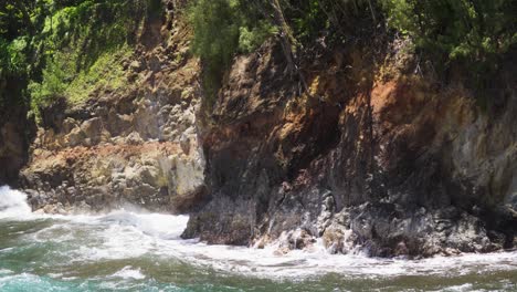 powerful surf rolls in and detonates against a rough cliff face, sending white spray up jagged rocks and along the shoreline, captured in bright daylight with tense, pounding rhythm