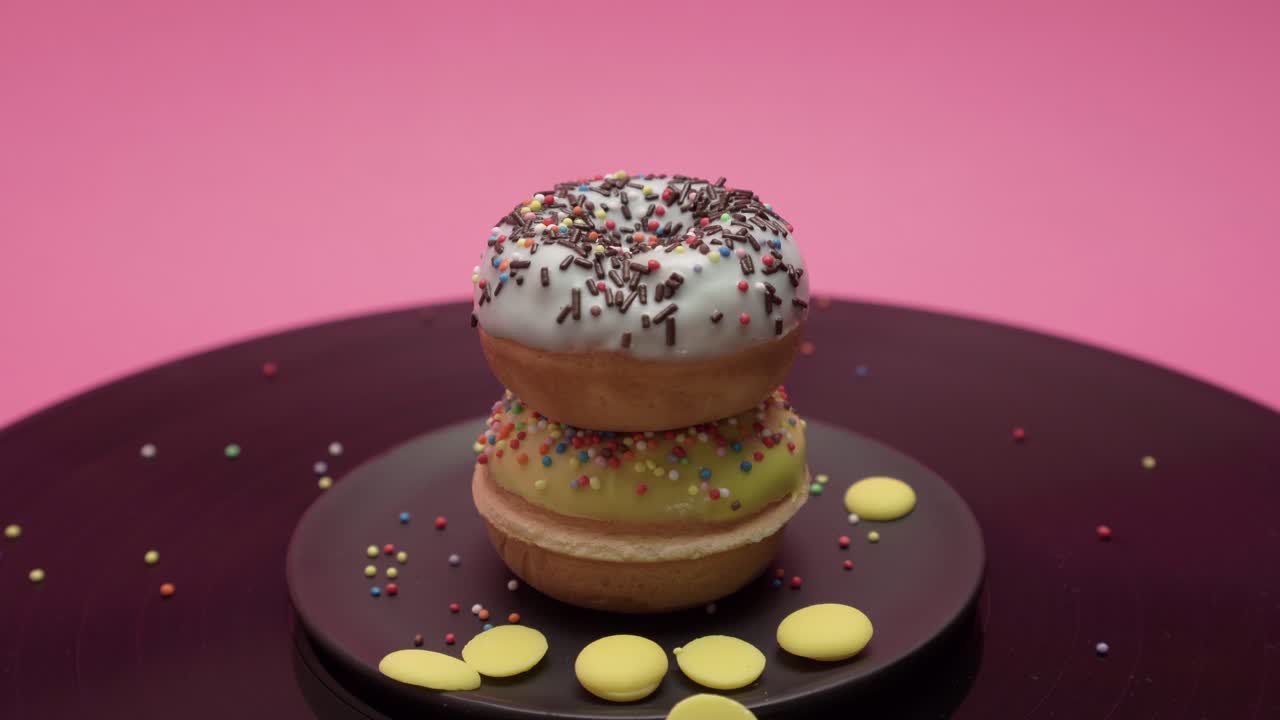 Two American donuts turn in circles on pink and black surfaces. Topped with yellow and blue chocolate with colorful crumbs