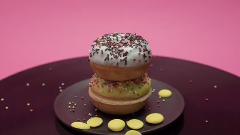 two american donuts turn in circles on pink and black surfaces. topped with yellow and blue chocolate with colorful crumbs