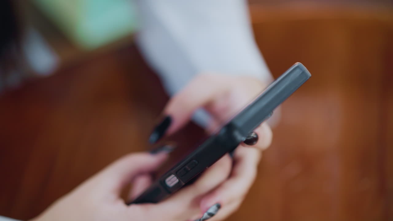 여성이 손을 칠한 검은색 전화의 클로즈업으로 전화로 메시지를 타이핑하고, 손은 무대를 둘러싸고 있는 부드러운 흐릿한 색의 배경으로 갈색 나무 테이블에 누워 있습니다.