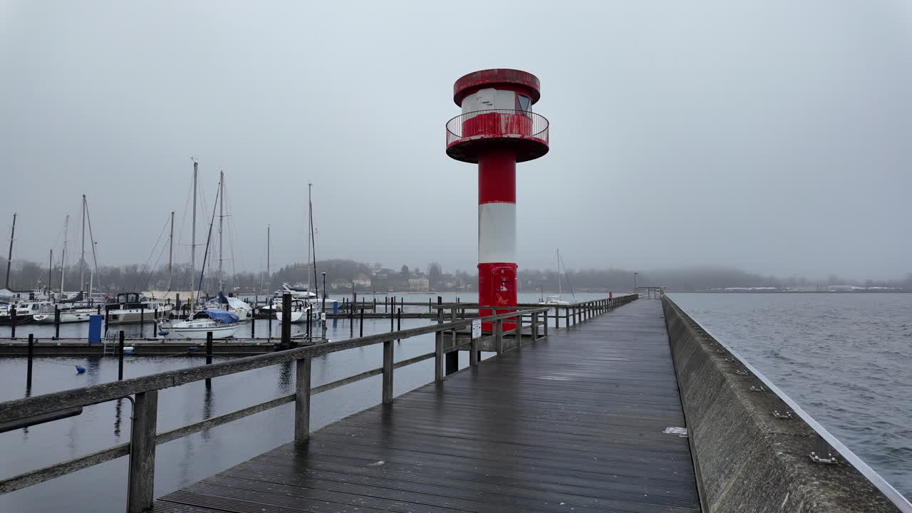 Vista panoramica del faro rosso e bianco all'ingresso del porto di Eckernförde, in Germania, immortalato in una giornata nebbiosa e nuvolosa. Un lungo molo di legno conduce verso il faro, con il Mar Baltico calmo