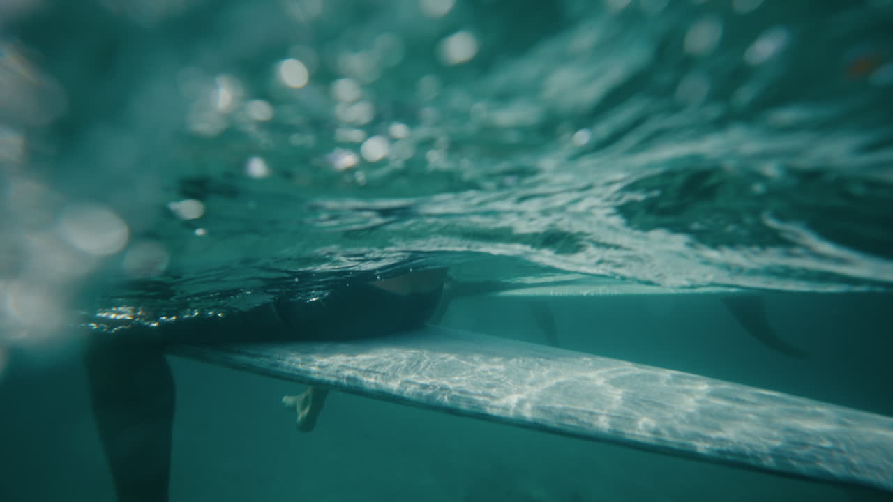 Ripresa subacquea metà fuori e metà sott'acqua di surfisti seduti in un oceano calmo e turchese vicino alla superficie.