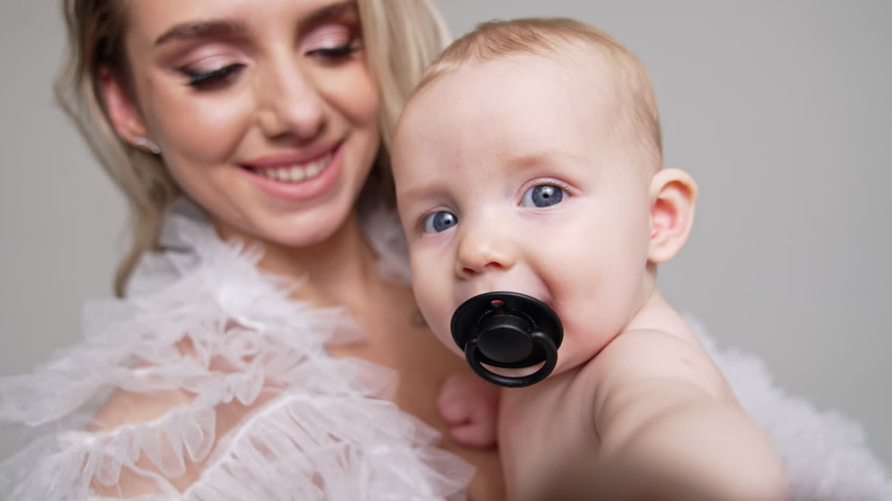 Adorable smiling Caucasian baby with grey eyes and pacifier in mouth trying to touch the camera. Mom and infant son close up portrait.