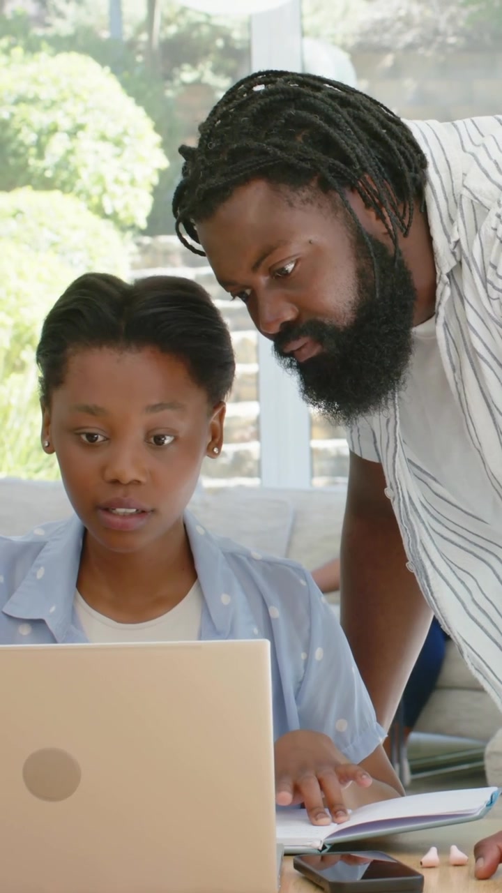 Vertical video: Approaching dad in striped tee leaning over child, guiding laptop and notes at home