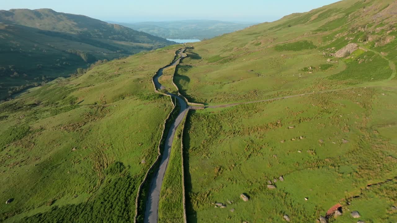 Wall lined mountainside road bathed in early morning sunshine heading towards distant lake Windermere. Summer. Kirkstone Pass, Lake District, Cumbria, UK