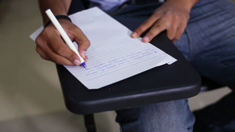 A-top-close-up-view-of-a-boy-student-writing-on-the-paper-at-his-class-room,-wearing-a-jeans-pant,-A-chair-with-its-writing-desk