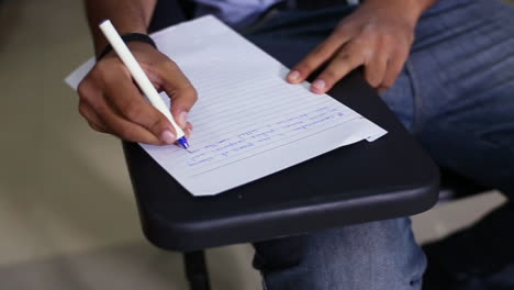 a top close up view of a boy student writing on the paper at his class room, wearing a jeans pant, a chair with its writing desk