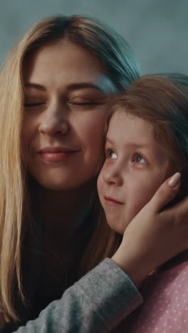 positive blonde mother hugs with adorable little daughter and cuddles spending time together in semi-dark room in evening at home close view slow motion