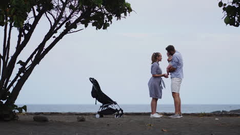 A-joyful-young-Caucasian-family,-accompanied-by-their-one-year-old-son-in-a-stroller,-strolls-along-a-sunlit-wooden-jetty.-The-scene-embodies-the-concept-of-a-happy-family