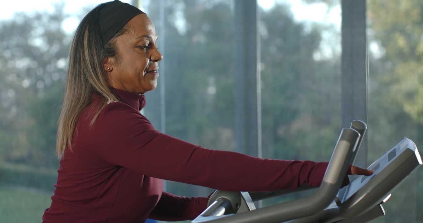 African American woman maroon top leaning, reaching console gripping bars, adjusting pace at gym