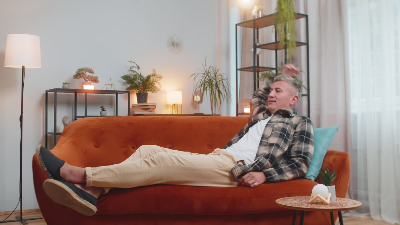 Tired young adult man sitting on comfortable couch taking a rest daytime nap resting at home room