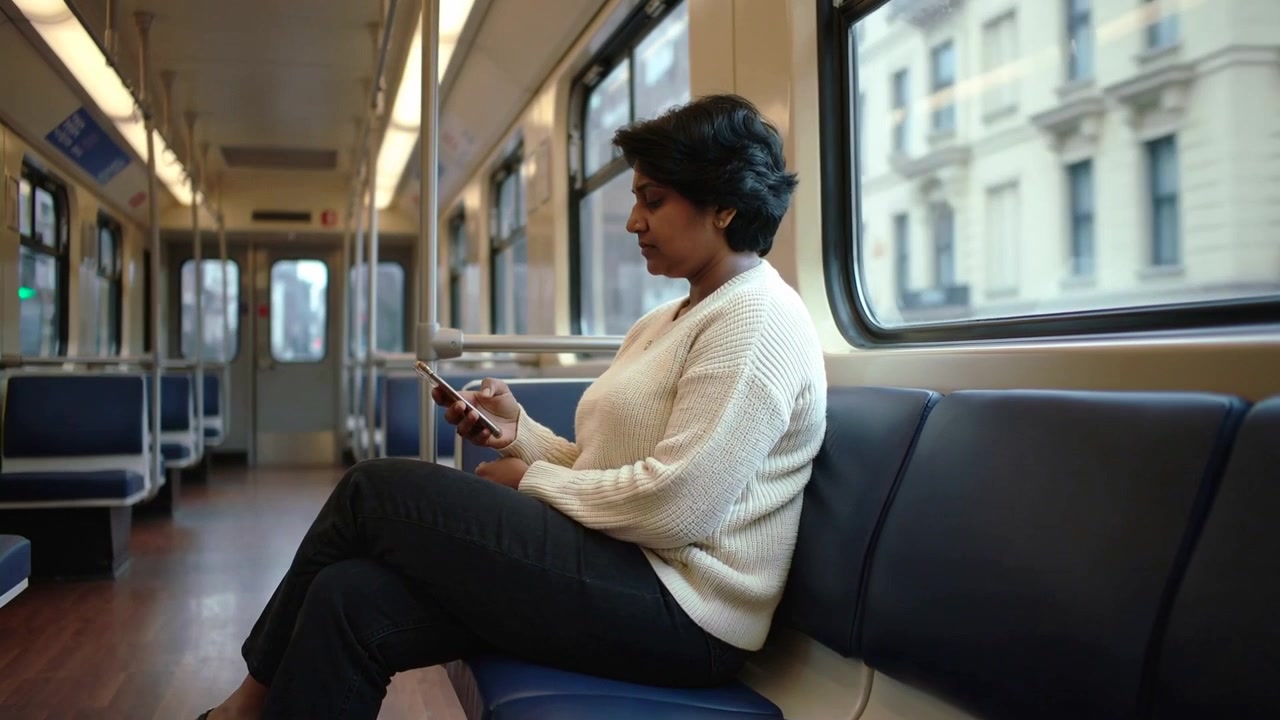 Scrolling Indian woman wearing cream sweater tapping phone in tram on blue bench while train moving
