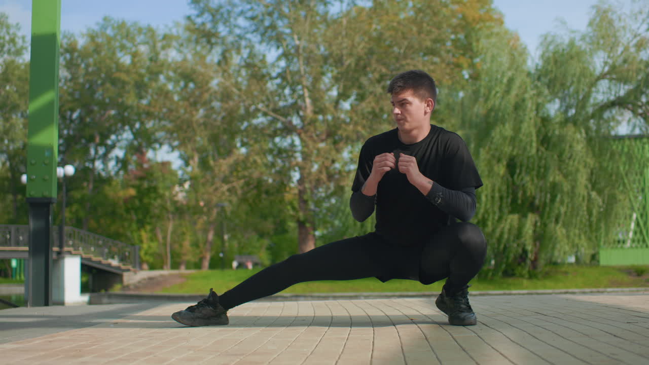 Uomo bianco che esegue un affondo laterale profondo vicino a un ponte e ad alberi. Un artista marziale a terra che allena l'equilibrio e il controllo. Un atleta in riabilitazione che si concentra su mobilità e forza. Respiri lenti e controllati. Sereno
