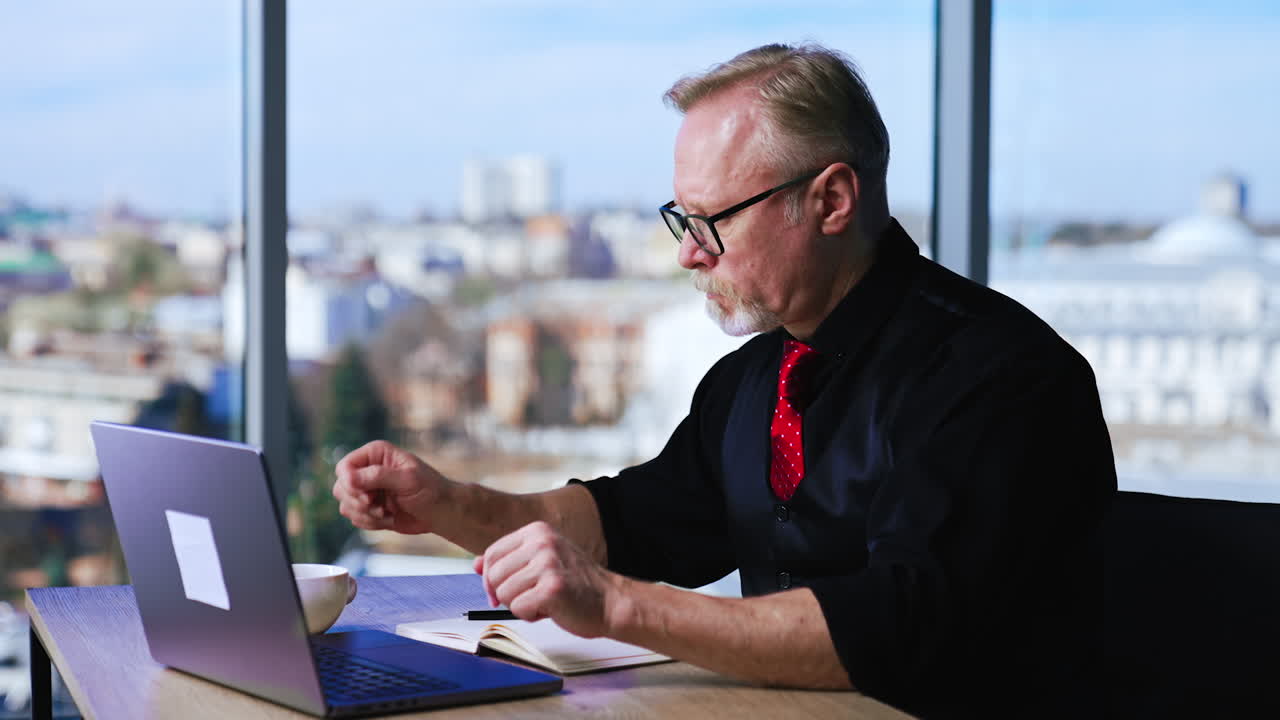 Career-oriented mature man types on his laptop in office. Boss looks at computer screen drinking coffee.