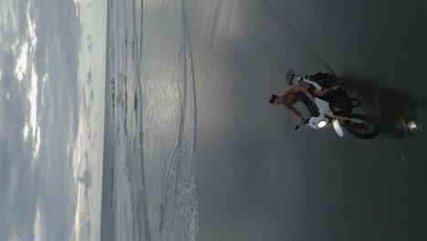 Cloudy-grey-vertical-aerial:-Asian-man-rides-motorcycle-on-beach