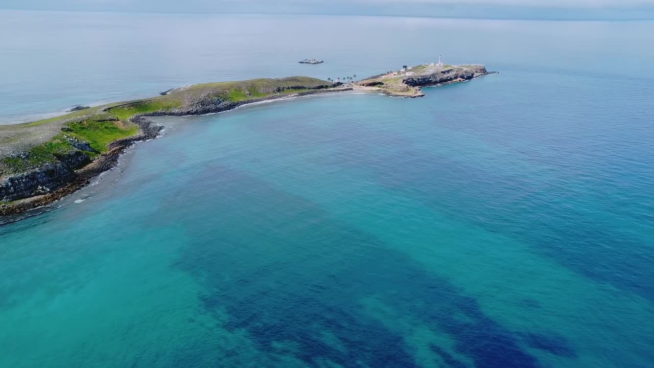 Isola dell'arcipelago di Abrolhos con un faro bianco che segna l'orizzonte, circondata da acque limpide turchesi e blu intenso dell'Oceano Atlantico al largo della costa di Bahia, Brasile
