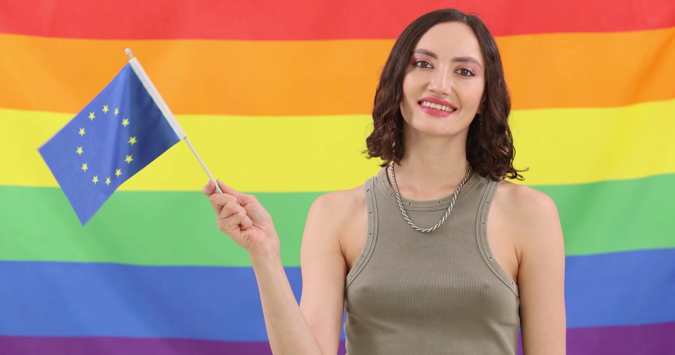 Donna sorride sventolando una piccola bandiera dell'Unione Europea davanti a uno striscione arcobaleno.
