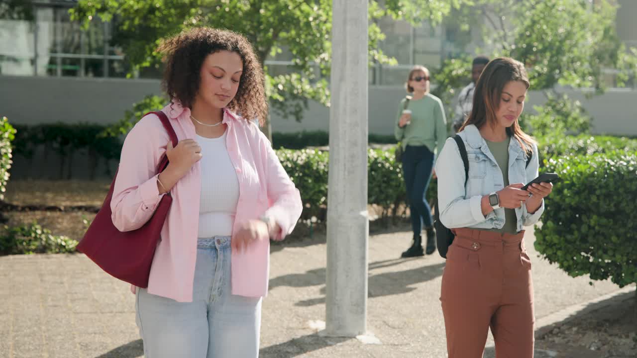 Donne afroamericane che aspettano su un sentiero, quella a sinistra che dà un'occhiata e sistema la tote bag, quella a destra che controlla il telefono vicino a un lampione