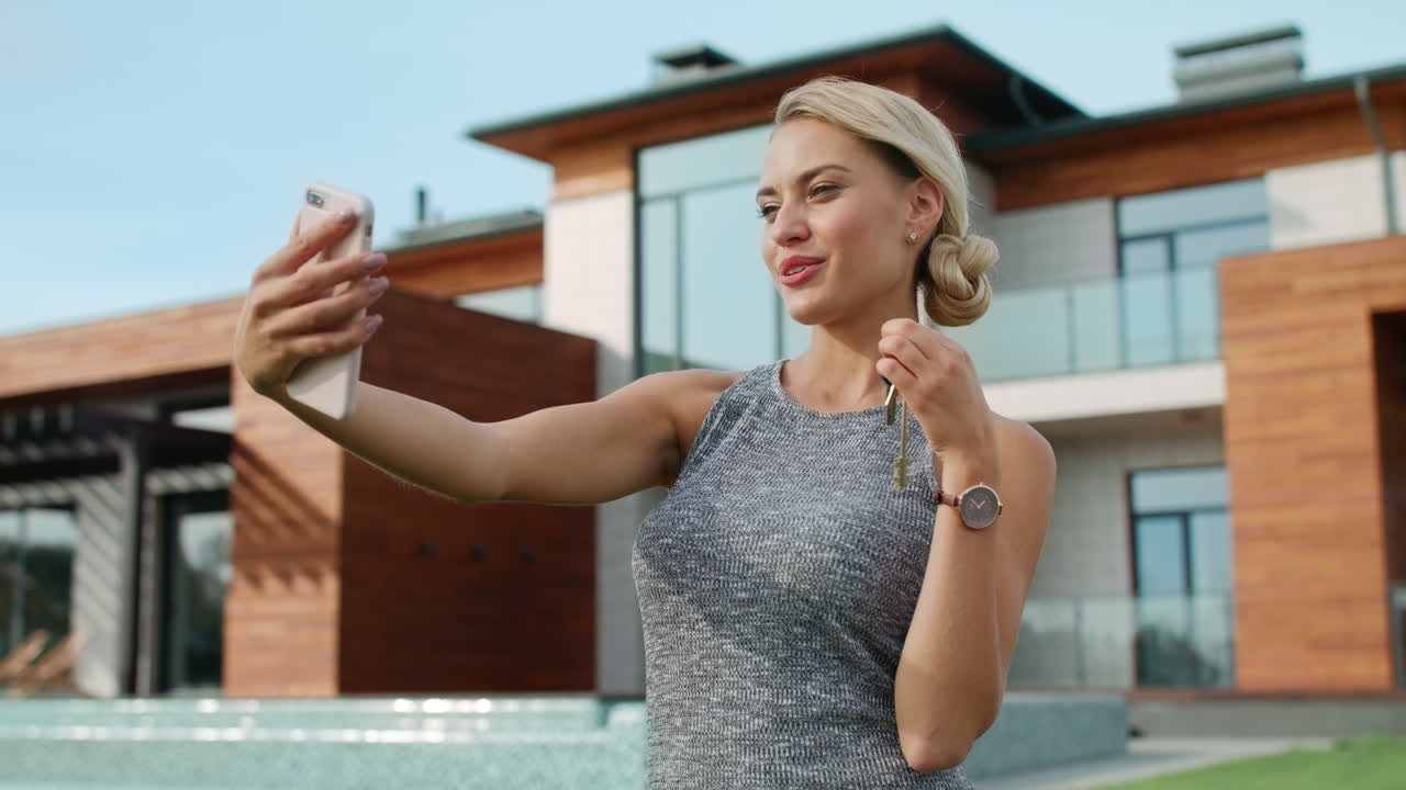 Happy woman taking selfie with house keys near luxury house.