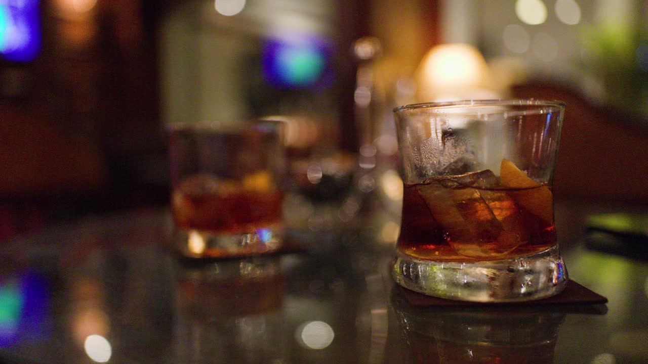 Hand picks up whiskey glass beside another drink in dimly lit, upscale bar setting