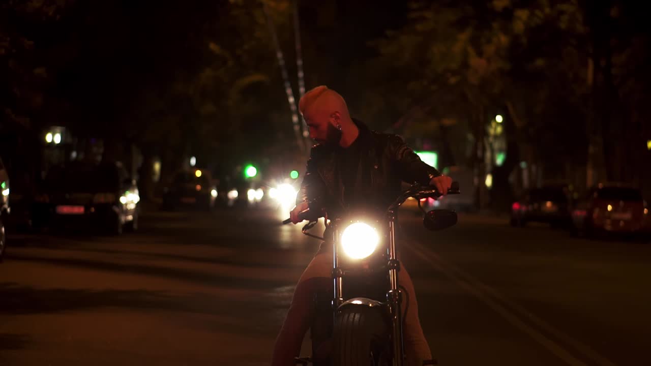 모터사이클에 앉아 있는 수염이 있는 라이더가 밤의 도시 거리에서 서 있는 헤드라이트를 밝게 돌리고 있습니다.