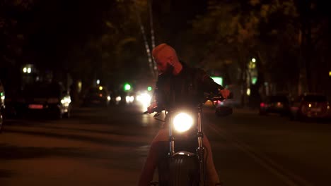 jinete barbudo sentado en una motocicleta, volviendo muy brillante el faro de pie en la calle de la ciudad de la noche